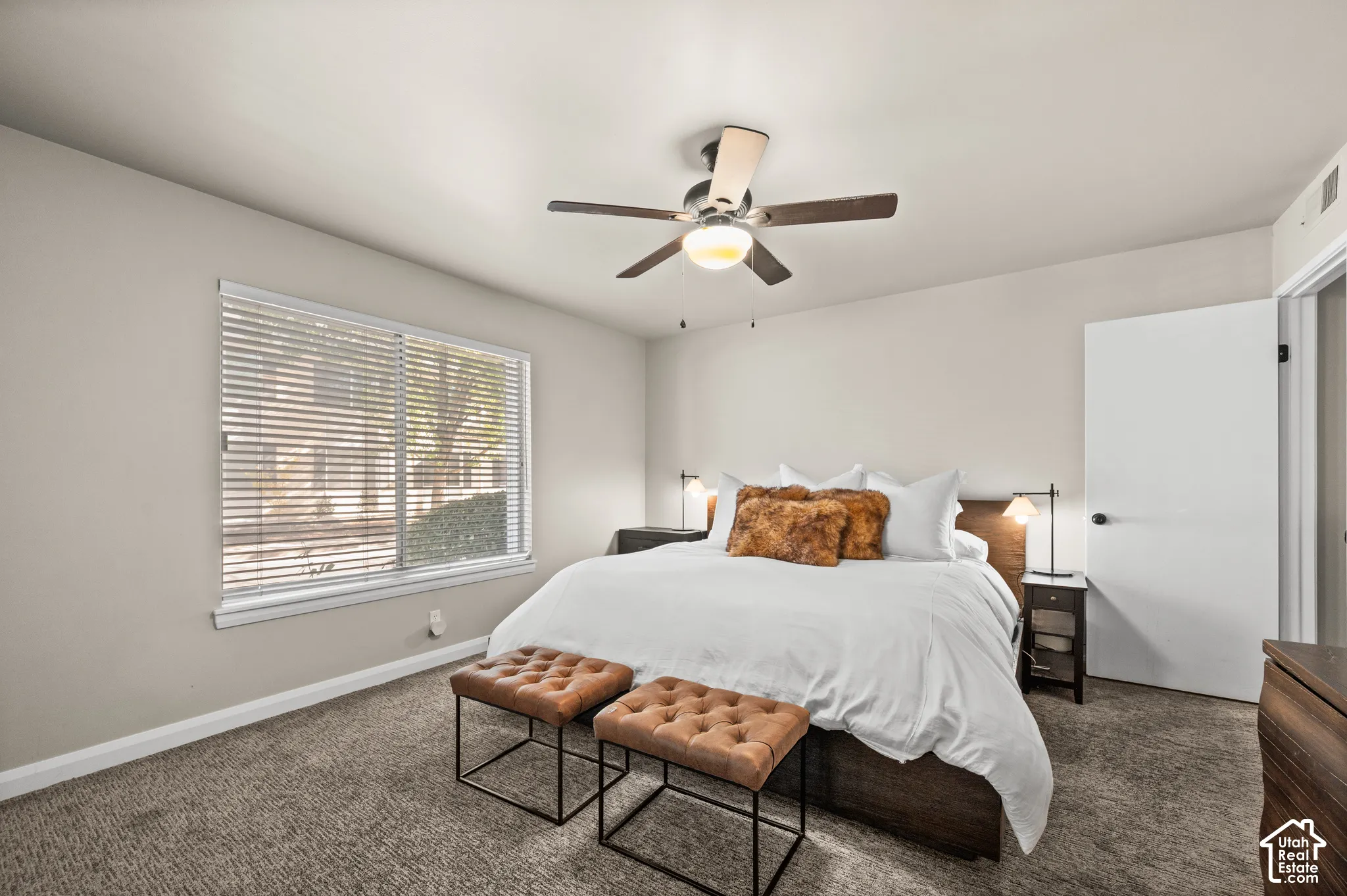 Neutral wall color, carpeted flooring, window with horizontal blinds, and a ceiling-mounted light fixture with a fan