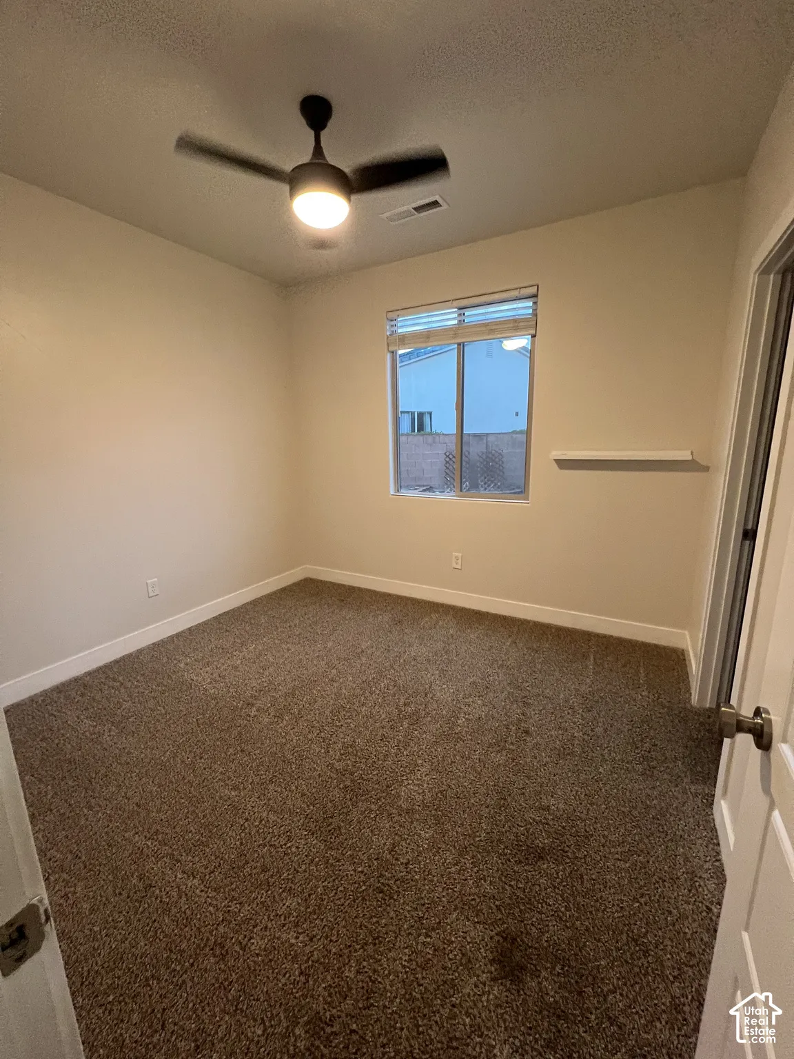 Neutral-toned room featuring wall-to-wall carpeting, a center-mounted ceiling fan with integrated lighting, a single-pane window with a horizontal blind, and a fixed wall-mounted shelf