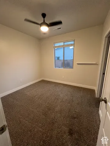 Neutral-toned room featuring wall-to-wall carpeting, a center-mounted ceiling fan with integrated lighting, a single-pane window with a horizontal blind, and a fixed wall-mounted shelf