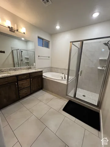 Bathroom featuring a dual-sink vanity with stone-finish countertops, tile flooring, a soaking tub with tile surround, and a glass-enclosed shower with integrated shelving