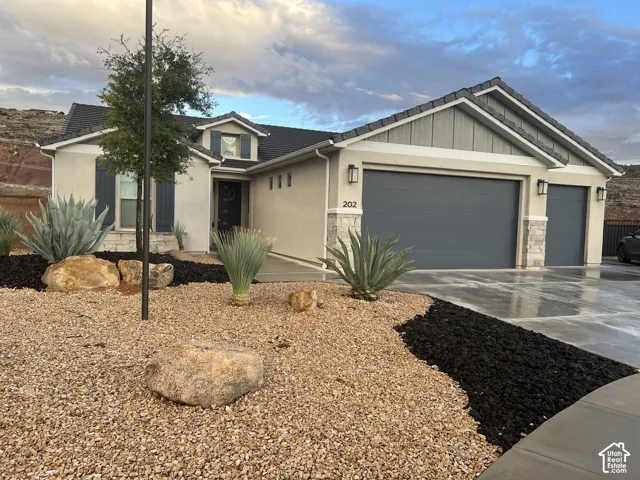 Stucco and stone veneer exterior featuring a three-car garage, vertical siding accents, and xeriscaped front yard with decorative rock and native vegetation