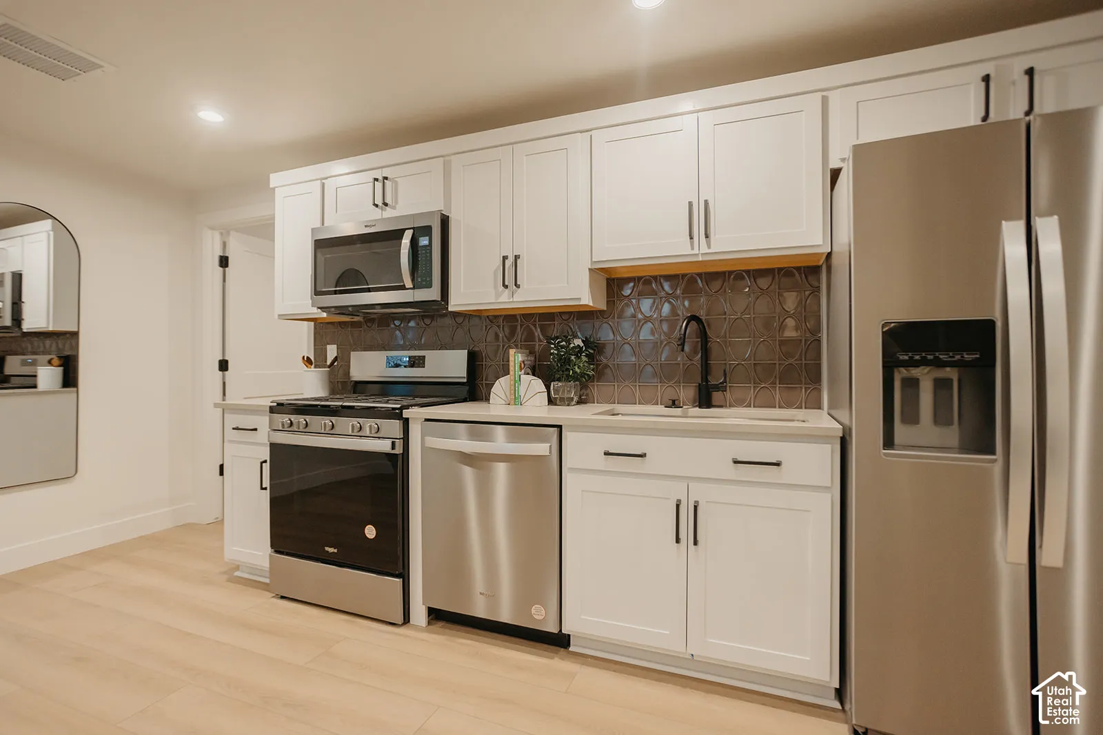 Kitchen featuring white shaker cabinetry, stainless steel appliances, patterned tile backsplash, wood-finish flooring, and a matte black gooseneck faucet