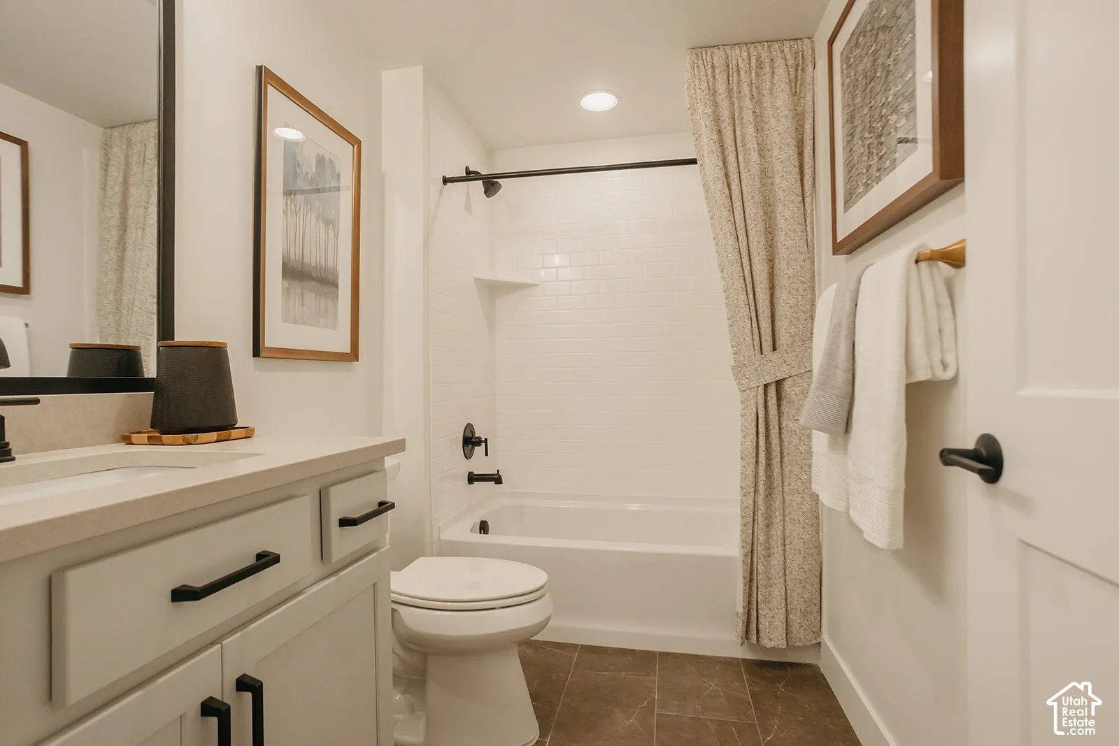 White vanity with a neutral solid-surface countertop, rectangular undermount sink, matte black hardware, subway-tiled tub surround, and porcelain tile flooring