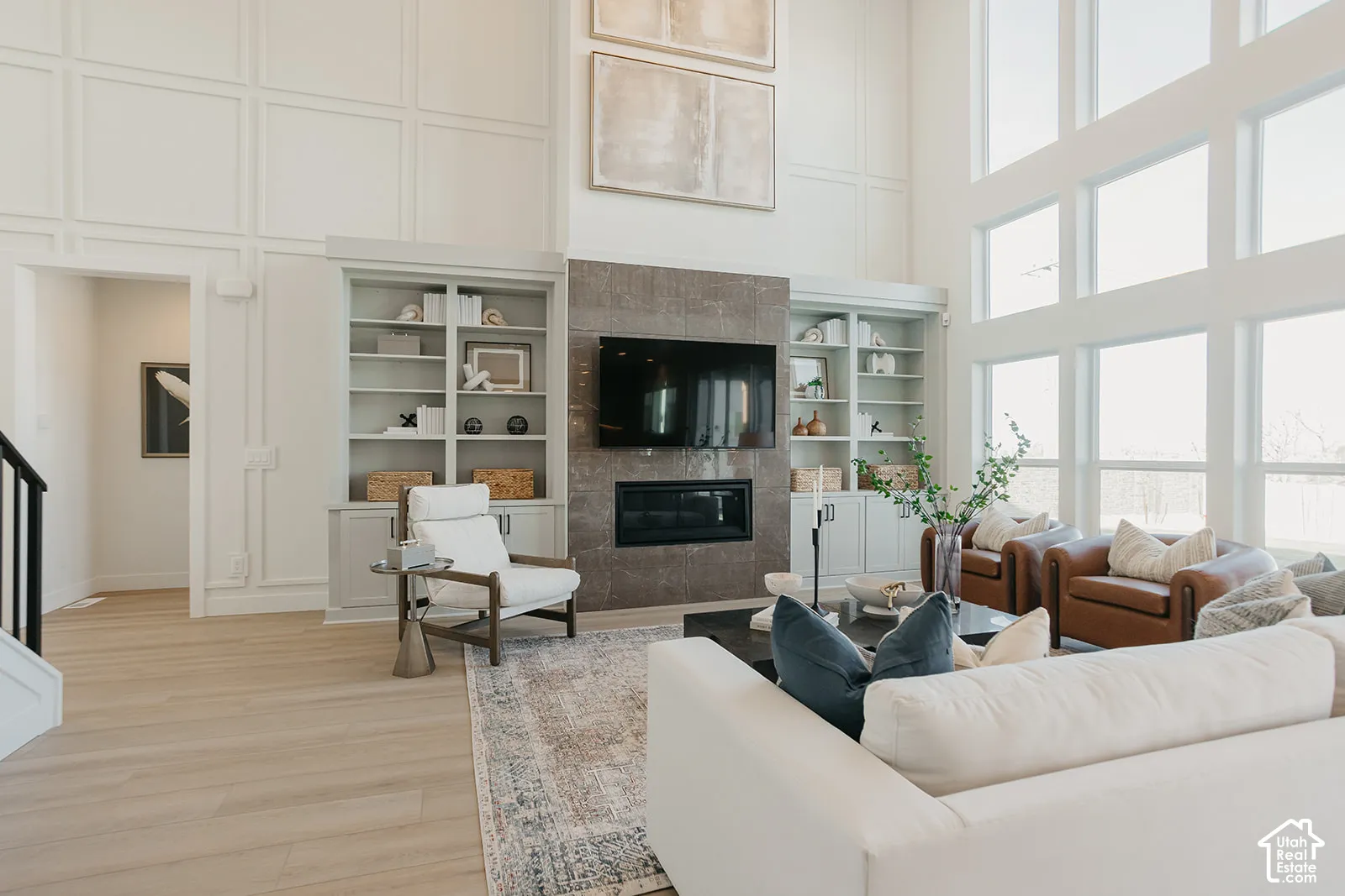 Double-height ceilings with floor-to-ceiling windows, custom built-in cabinetry, wood-finish flooring, and a fireplace with stone surround