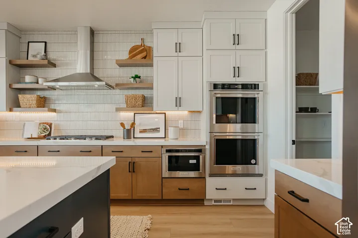 Kitchen featuring two-tone cabinetry, stainless steel double wall ovens, a stainless steel range hood, stone-finish countertops, and linear tile backsplash