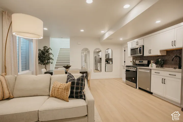 Open-concept kitchen featuring white shaker-style cabinetry, stainless steel appliances, dark tile backsplash, and wood-finish flooring with recessed lighting