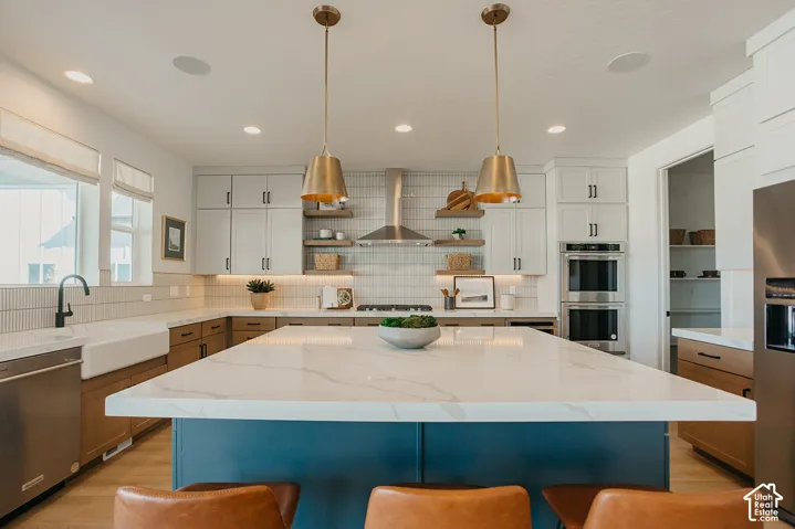 Kitchen featuring a large central island with a stone-surface countertop, white shaker-style cabinetry, stainless steel double wall ovens, a farmhouse sink with a matte black faucet, and metallic pendant lighting