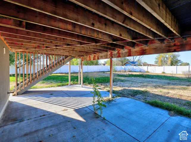 Covered concrete patio featuring exposed wood beam joist ceiling, integrated exterior staircase, and vinyl perimeter fencing