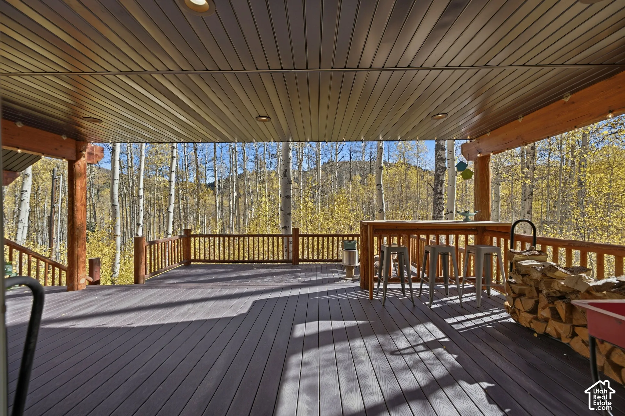 Covered deck featuring composite decking, recessed lighting, exposed timber beams, and wood railing overlooking a forested landscape