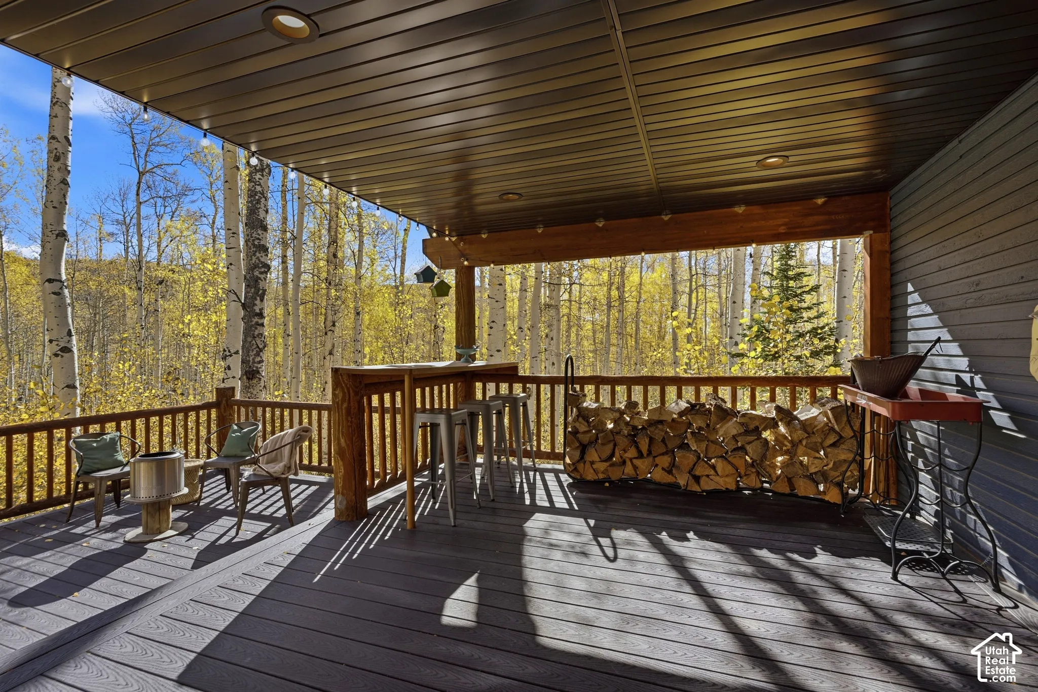 Covered outdoor deck featuring composite decking, built-in wooden bar, recessed lighting, and wooden railing overlooking a wooded forest