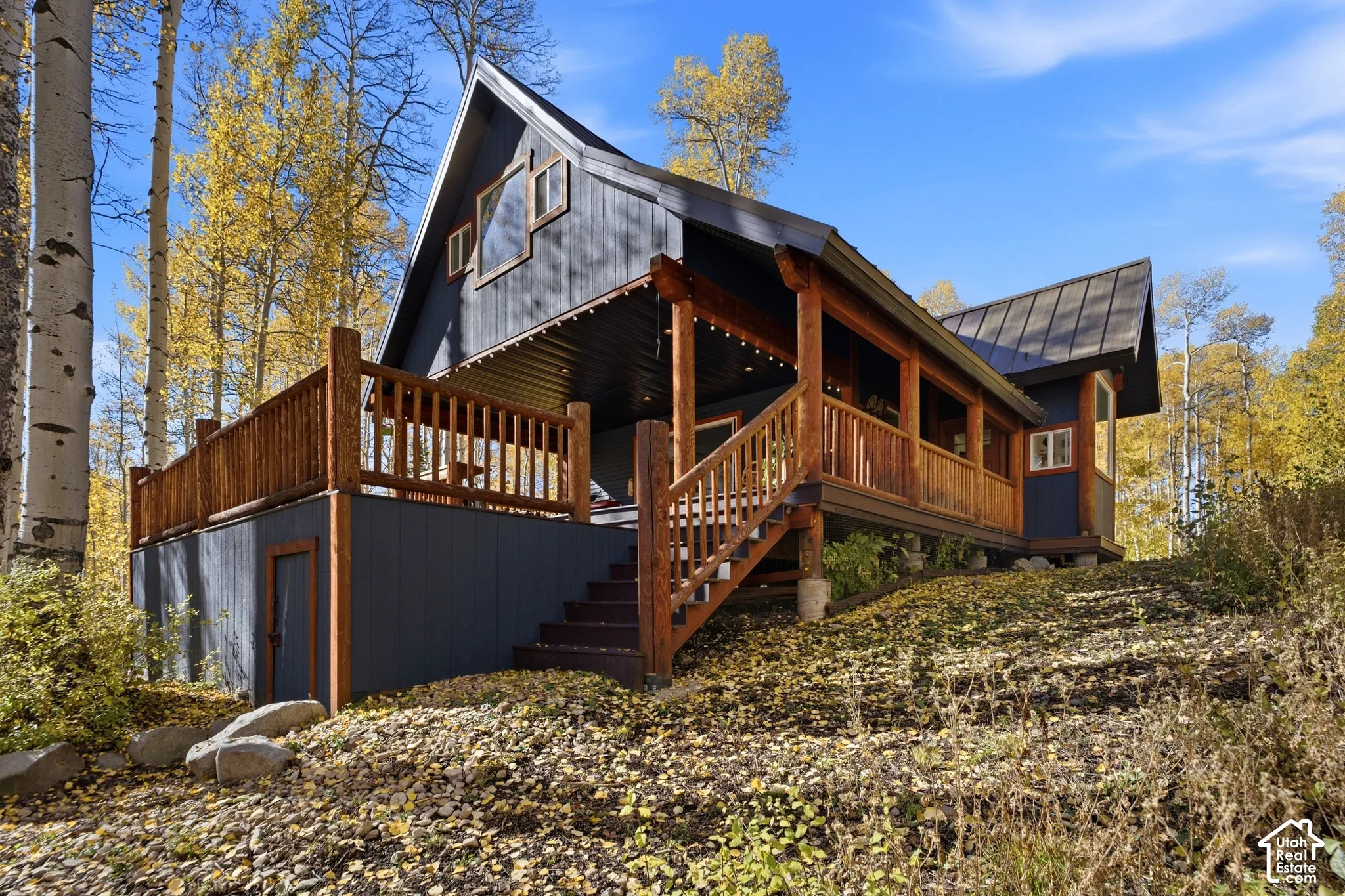 Architectural cabin exterior featuring vertical dark-tone siding, a metal roof, prominent log timber support beams, and a wrap-around wood deck with integrated railings