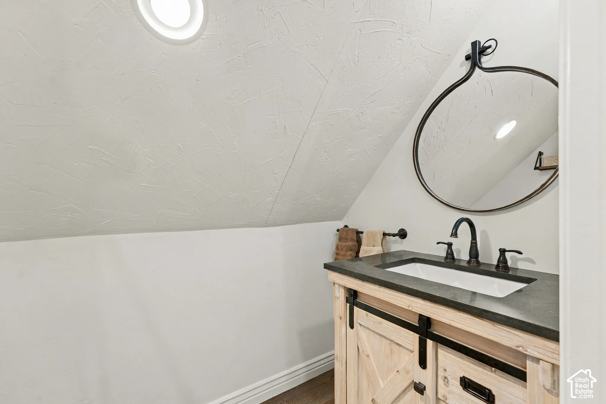 Wood-finish vanity with sliding barn-style doors, stone-composite countertop, rectangular undermount sink, dark bronze faucet, and circular wall-mounted mirror