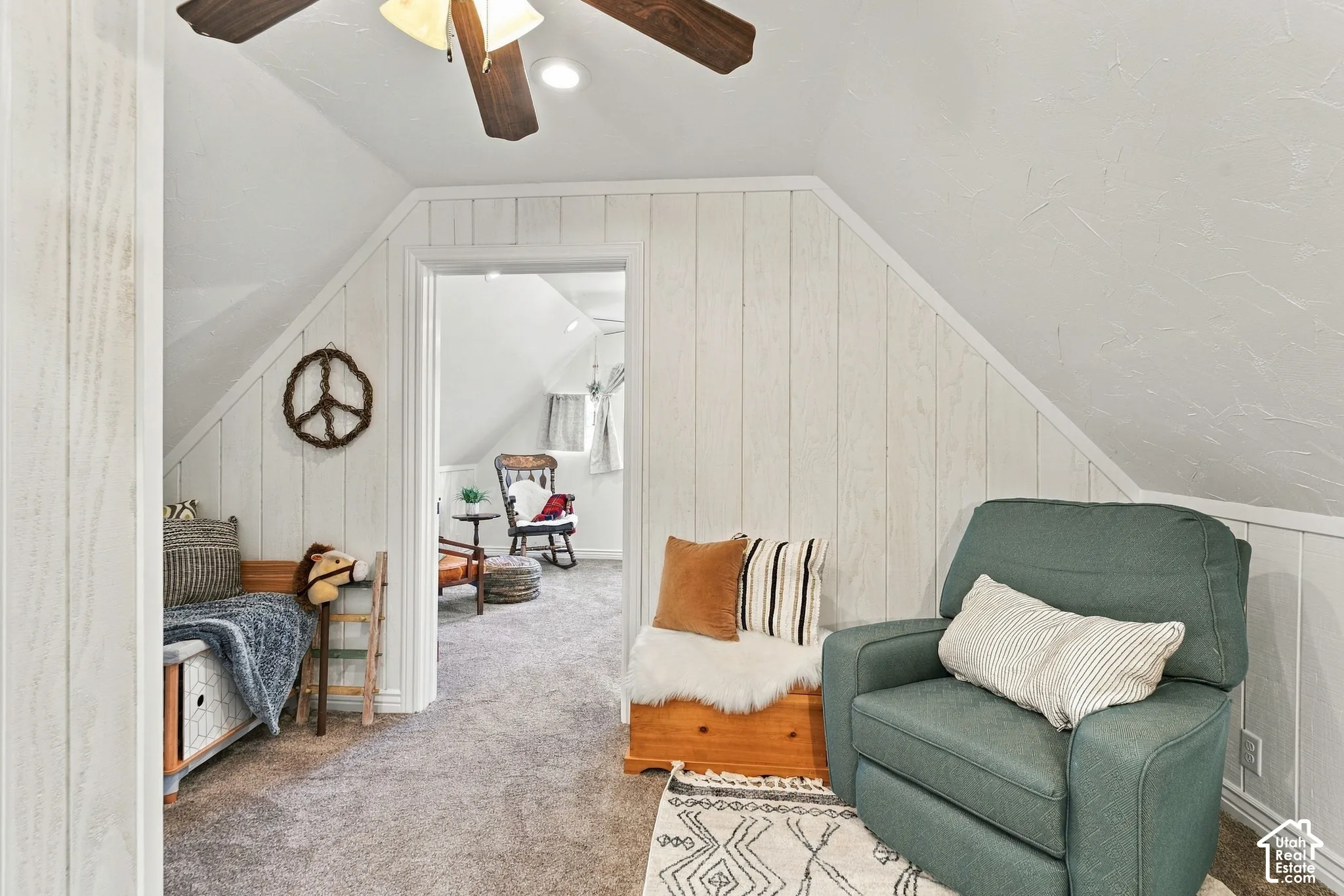 Vaulted ceilings featuring vertical white-painted wood wall paneling, neutral carpeting, and a ceiling fan with light fixture