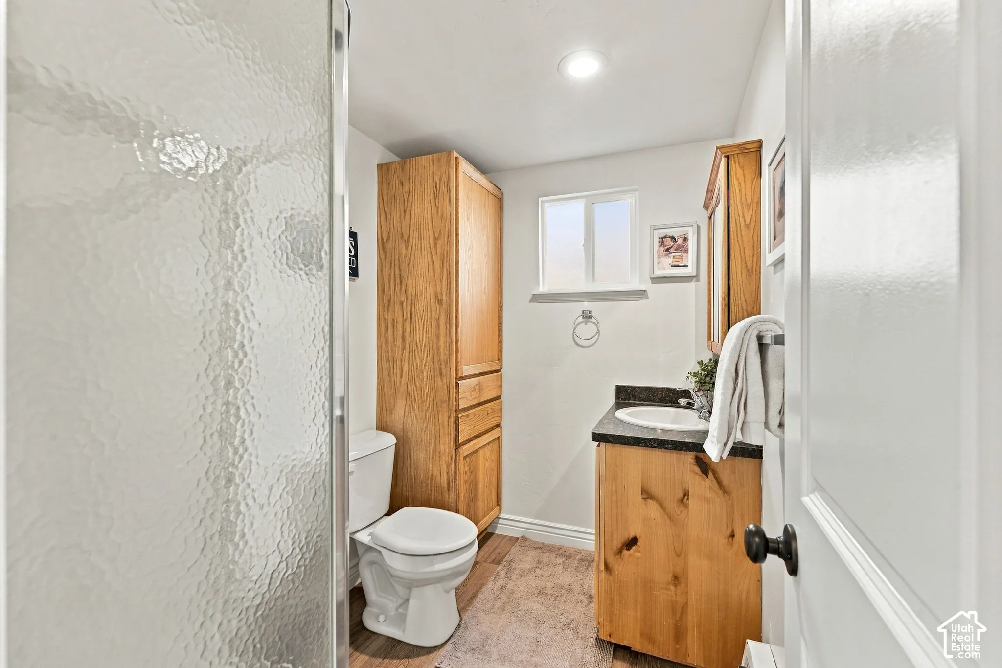 Bathroom featuring wood-finish cabinetry, a built-in floor-to-ceiling storage cabinet, dark stone-style countertop with an integrated basin, and a glass-enclosed shower