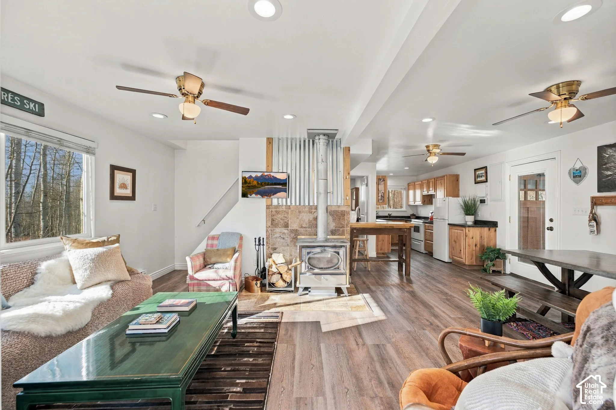 Open-concept area featuring wood-finish flooring, a wood-burning stove with stone surround and corrugated metal accent wall, recessed lighting, and ceiling fans