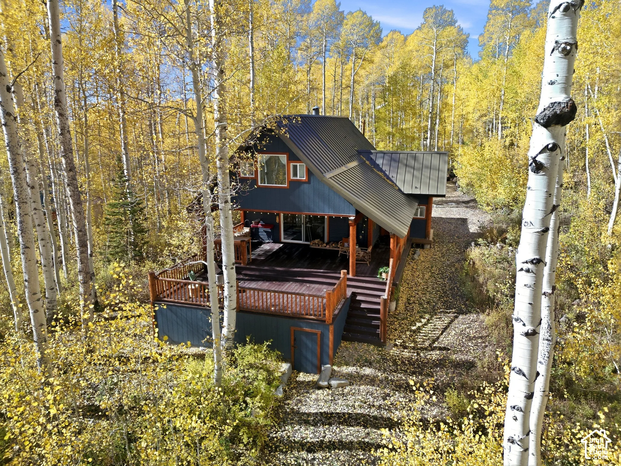 Two-story residence featuring a dark blue exterior, metal roof, wrap-around wooden deck with railings, and surrounded by mature aspen trees