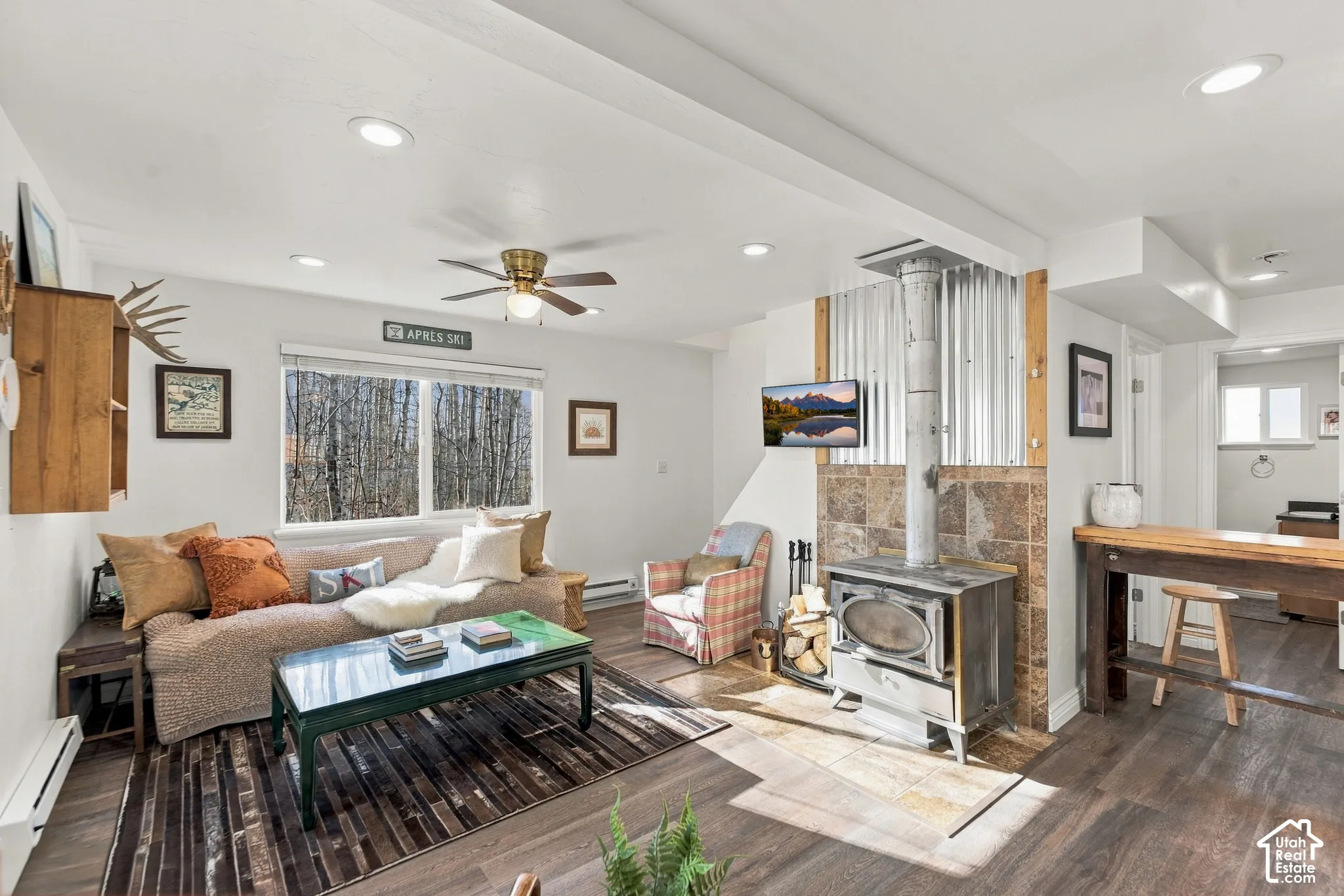 Living space featuring wood-finish flooring, recessed lighting, ceiling fan, and a wood-burning stove with stone surround and corrugated metal accent wall