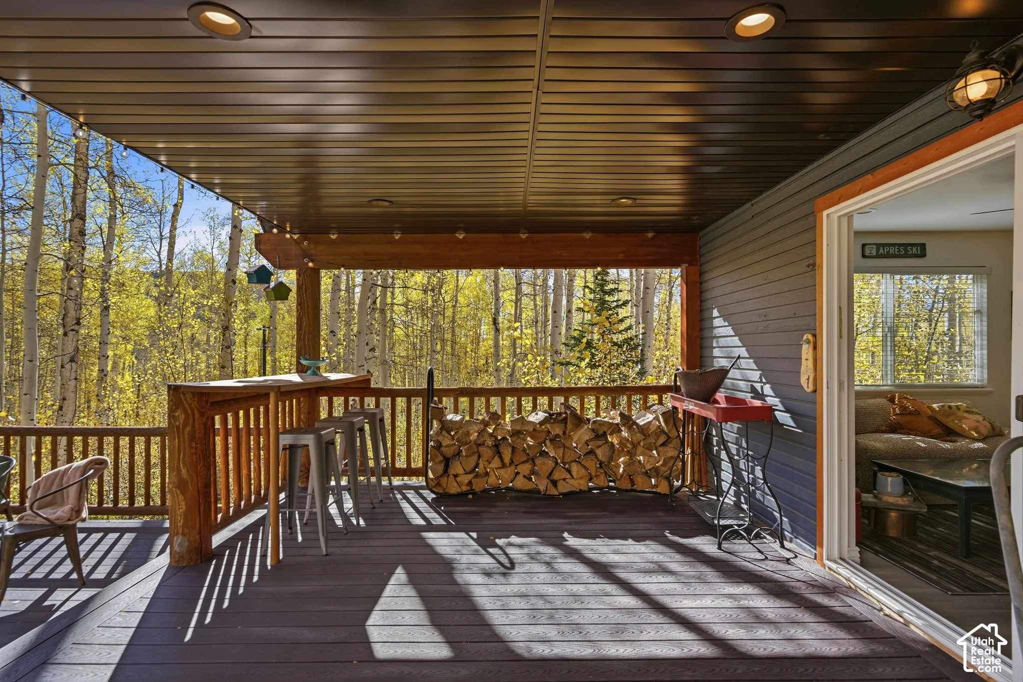 Covered deck featuring composite decking, built-in wooden bar, integrated recessed lighting, and wooden railing overlooking a wooded area