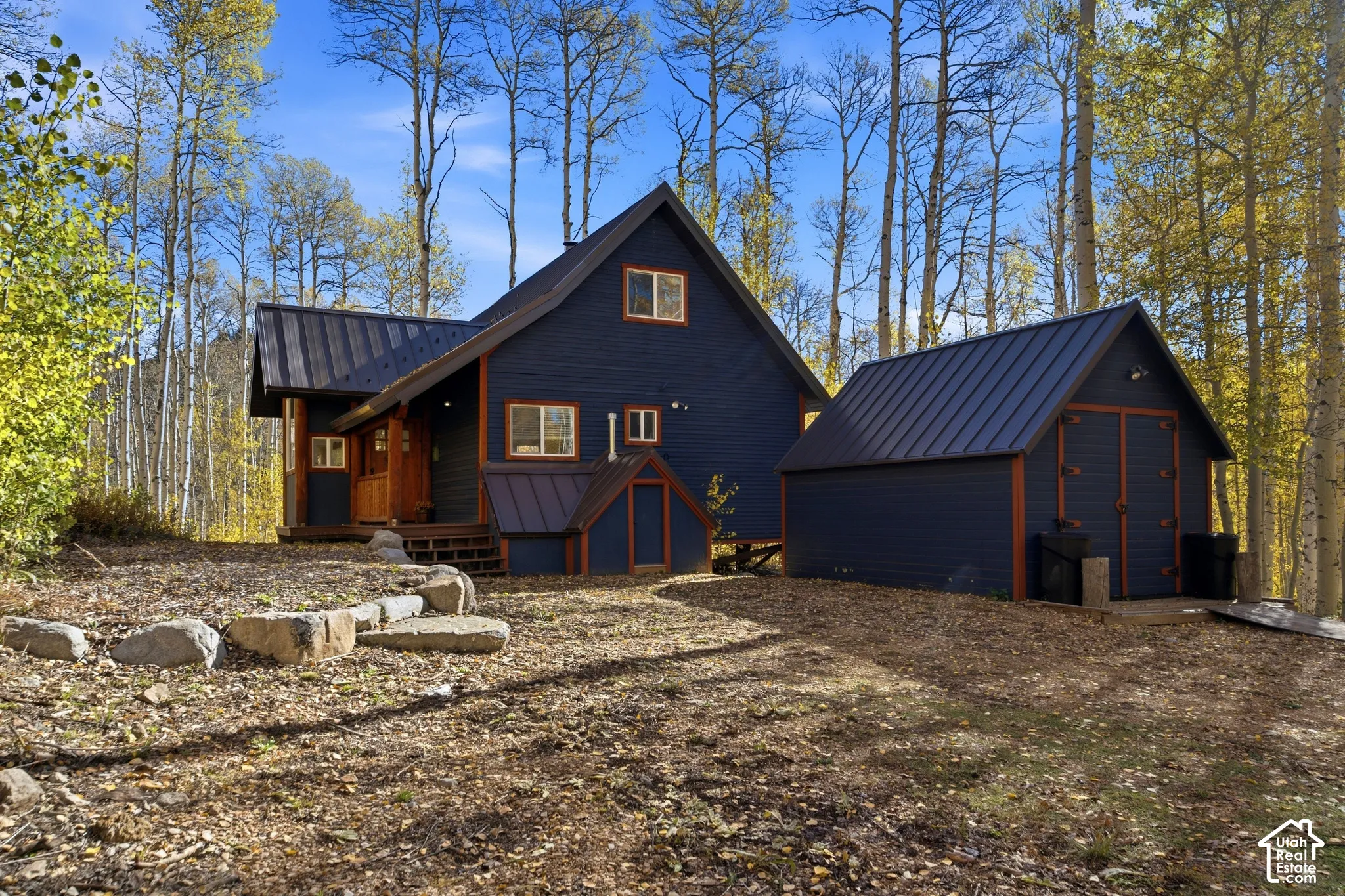Dark-stained horizontal siding with timber trim, charcoal-colored metal roofing, and a detached structure situated among mature aspen trees