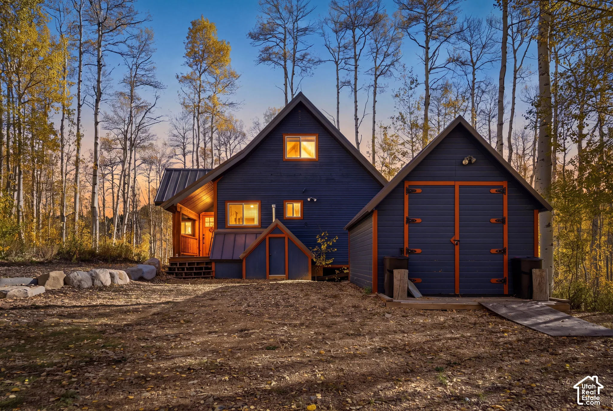 Two-story cabin exterior featuring painted wood siding, metal roofing, a detached structure with dual swinging doors, and surrounding mature aspen trees