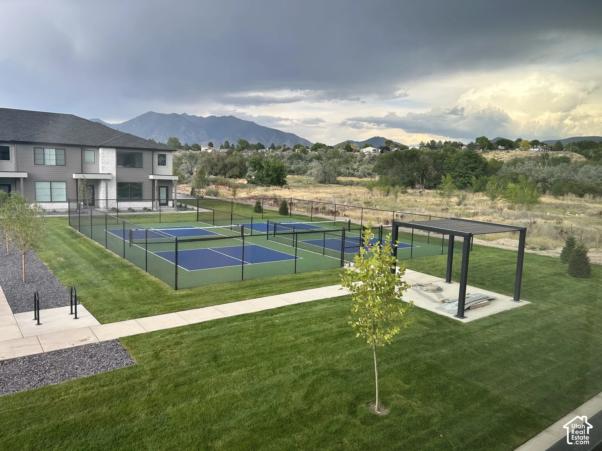 Fenced pickleball courts with blue surfacing, integrated metal pergola, manicured lawn, and mountain backdrop