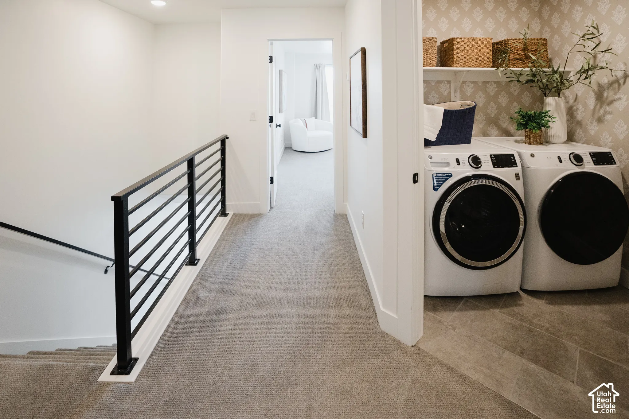 Laundry space featuring tile flooring, patterned wallpaper, and built-in shelving, complemented by a staircase with a matte black metal railing
