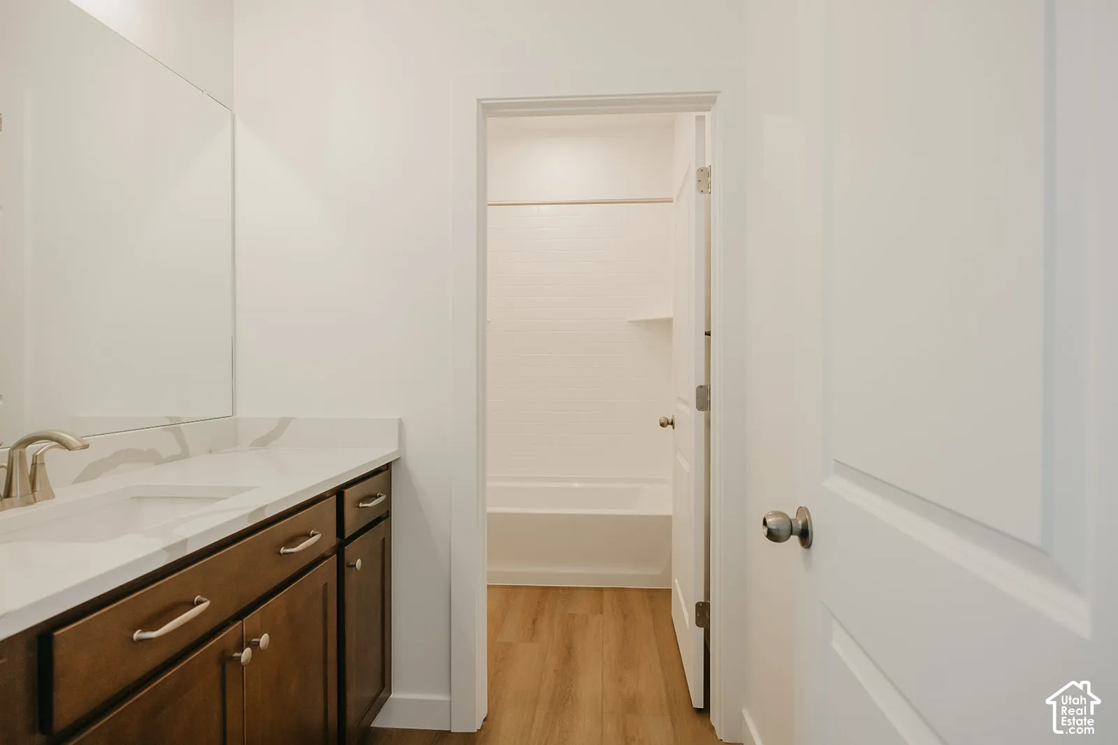 Bathroom featuring a vanity with wood-finish cabinetry, a stone-composite countertop with an integrated sink, a large wall-mounted mirror, and wood-finish flooring extending into the tub area