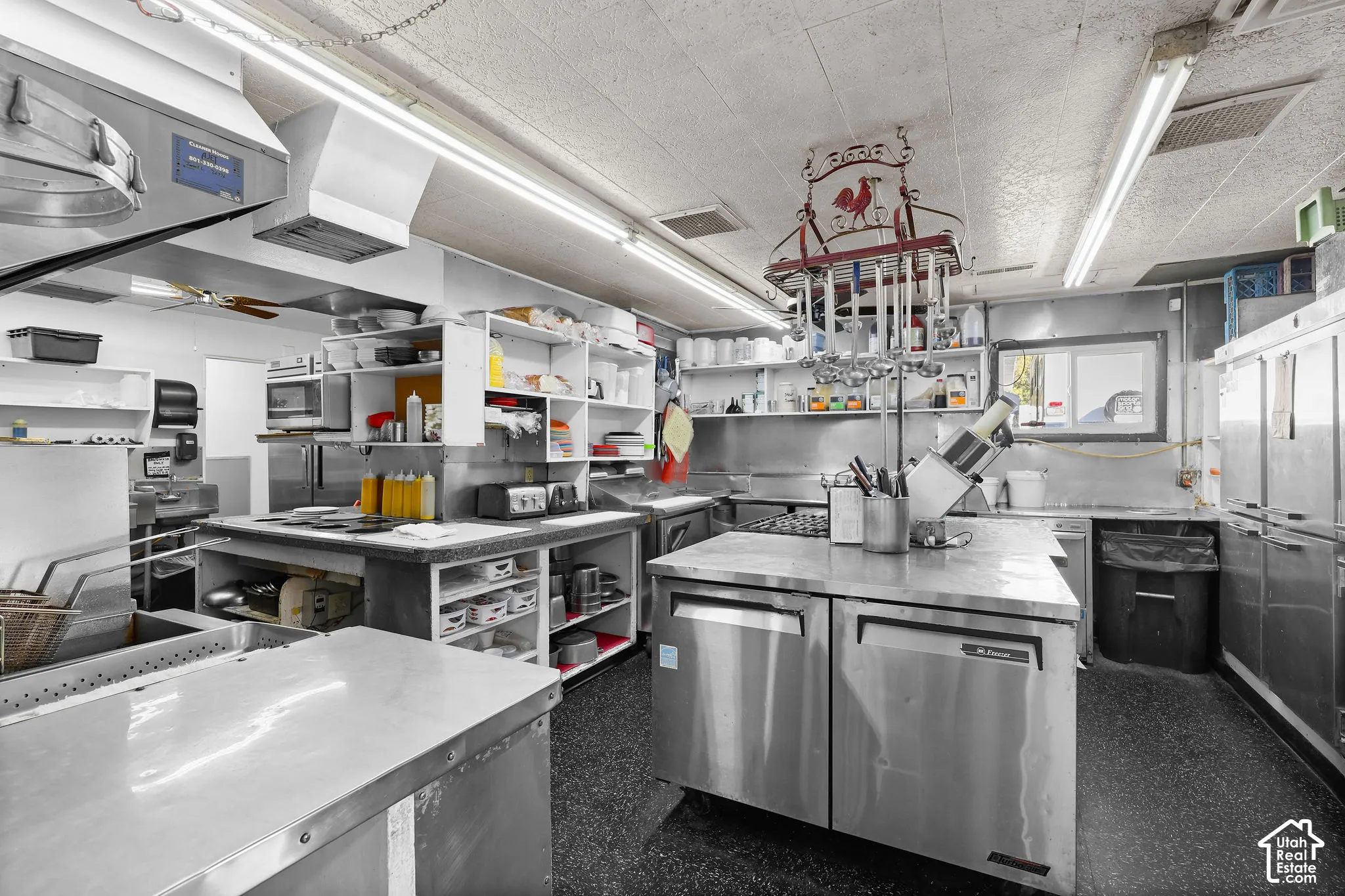 Commercial kitchen space featuring stainless steel work surfaces, heavy-duty stainless steel refrigeration units, a stainless steel exhaust hood, open industrial shelving, and high-output linear lighting
