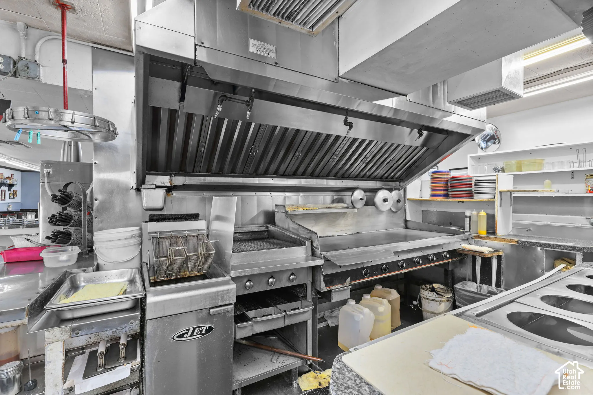 Commercial kitchen workspace featuring a stainless steel ventilation hood, industrial flat-top grill, deep fryer, and built-in prep surfaces