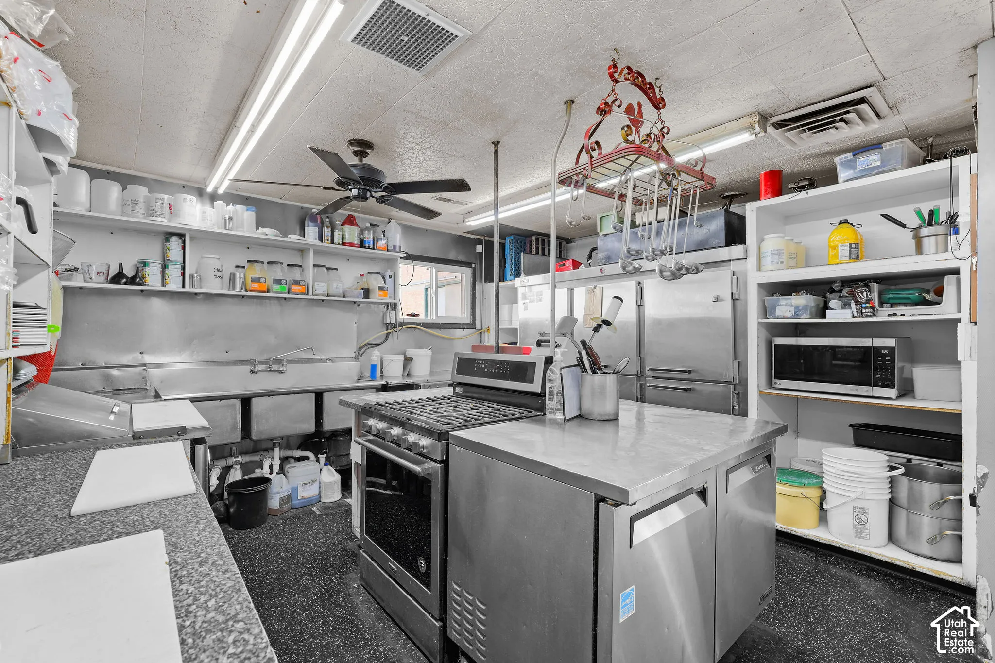 Commercial-grade kitchen featuring stainless steel prep stations, a multi-basin sink, gas range, metal shelving, and a ceiling-mounted rack