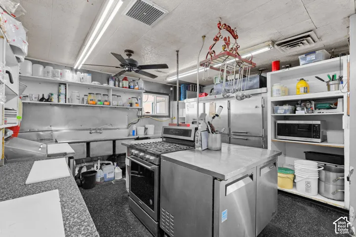 Commercial-grade kitchen featuring stainless steel prep stations, a multi-basin sink, gas range, metal shelving, and a ceiling-mounted rack