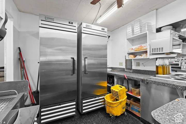Commercial kitchen space featuring two stainless steel upright refrigerators, stone-finish countertops, wall-mounted shelving, and overhead fluorescent lighting