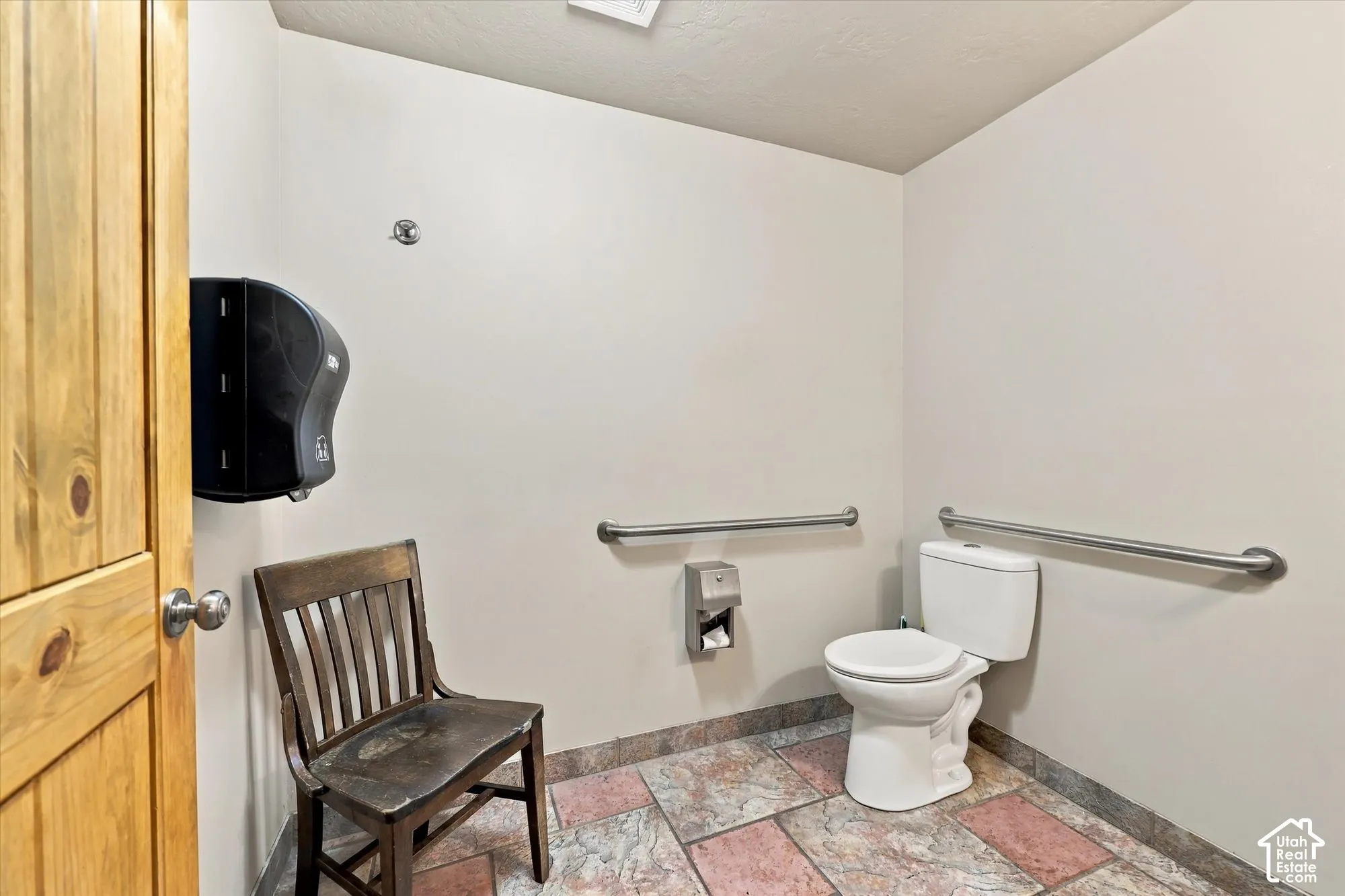 Restroom featuring multi-tonal tile flooring, stainless steel grab bars, a dual-flush toilet, and a wall-mounted commercial paper dispenser