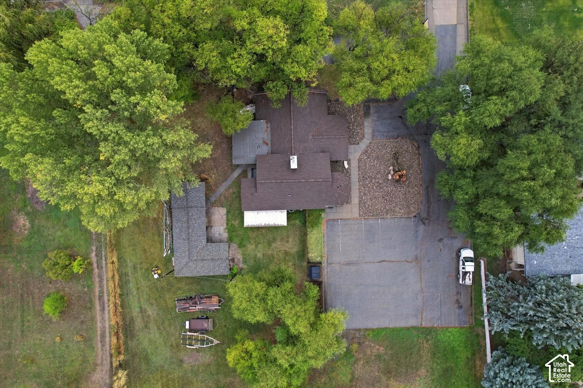 Aerial view featuring a multi-section roofline, expansive paved driveway, stone-surfaced exterior area, and surrounding mature trees