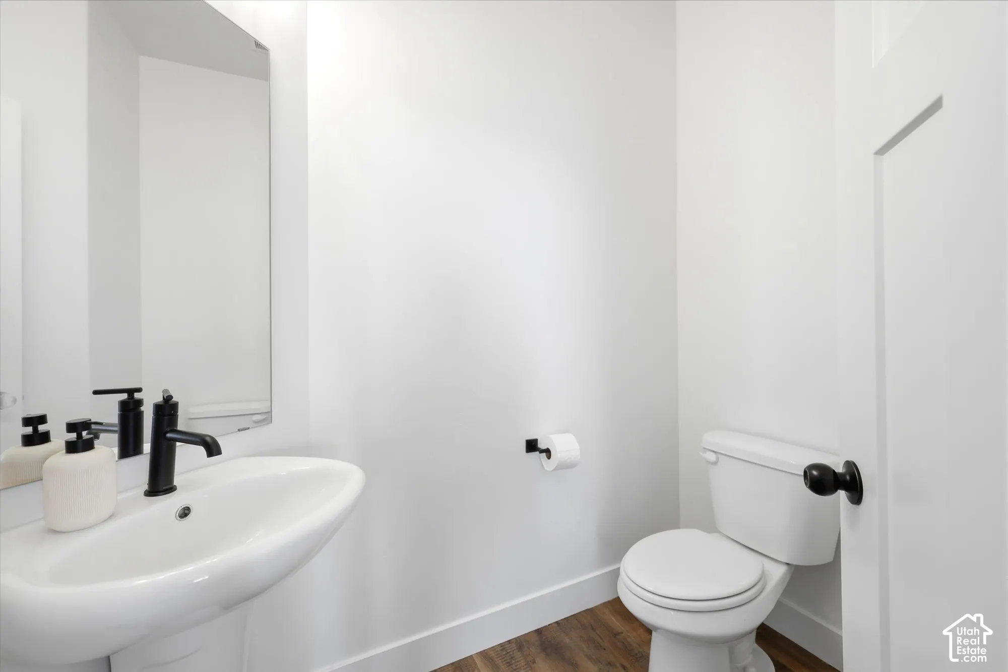Powder room featuring a pedestal sink, matte black faucet, wall-mounted mirror, and wood-finish flooring