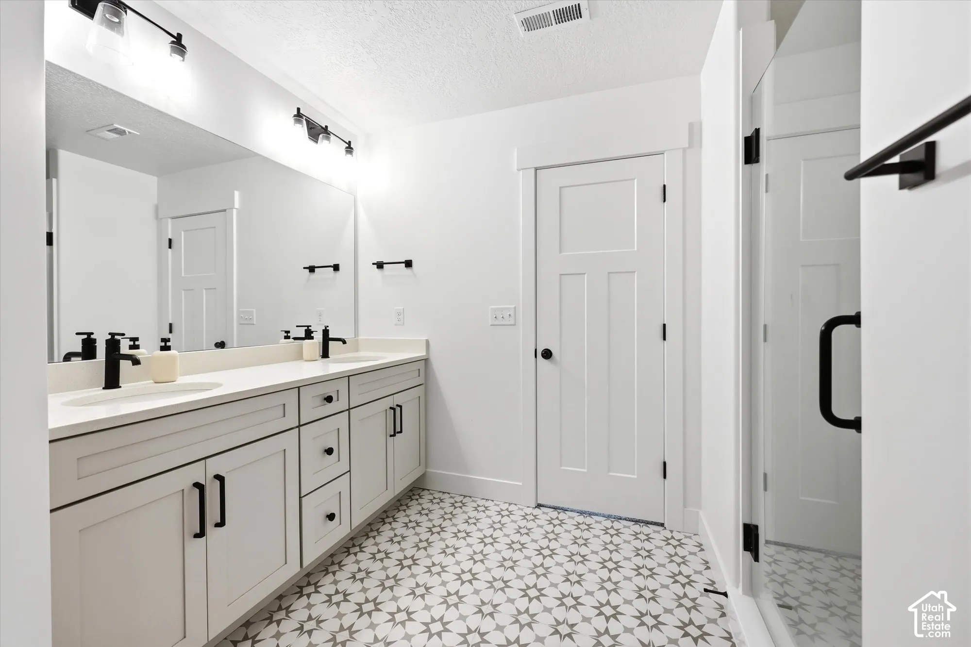 Dual-sink vanity with matte black fixtures, quartz countertop, patterned tile flooring, and glass-enclosed shower with matte black hardware
