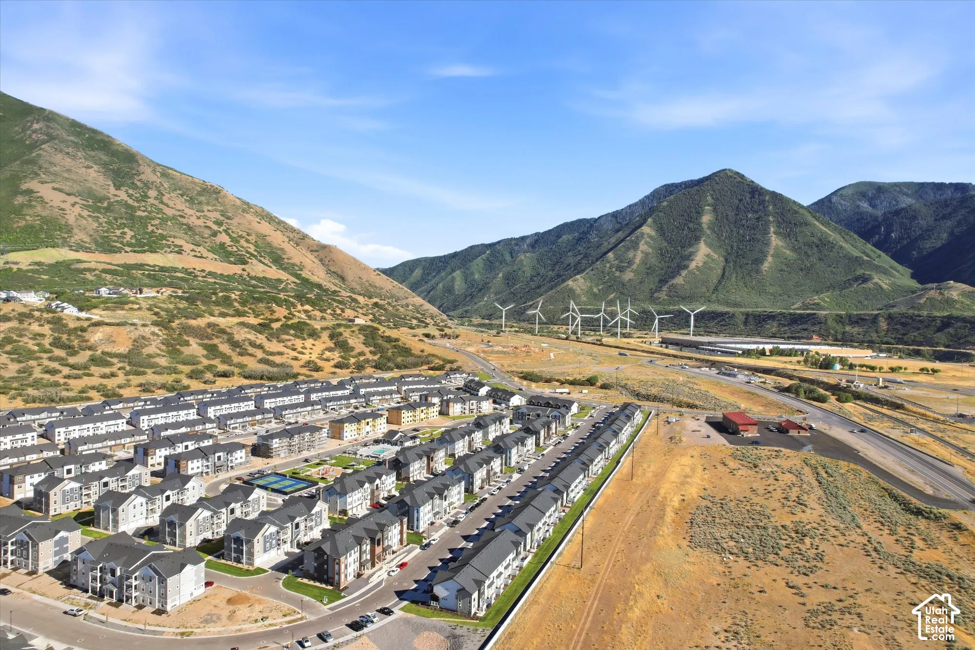 Aerial perspective of a multi-building residential development featuring mountain scenery, paved streets, exterior parking areas, and on-site recreational courts