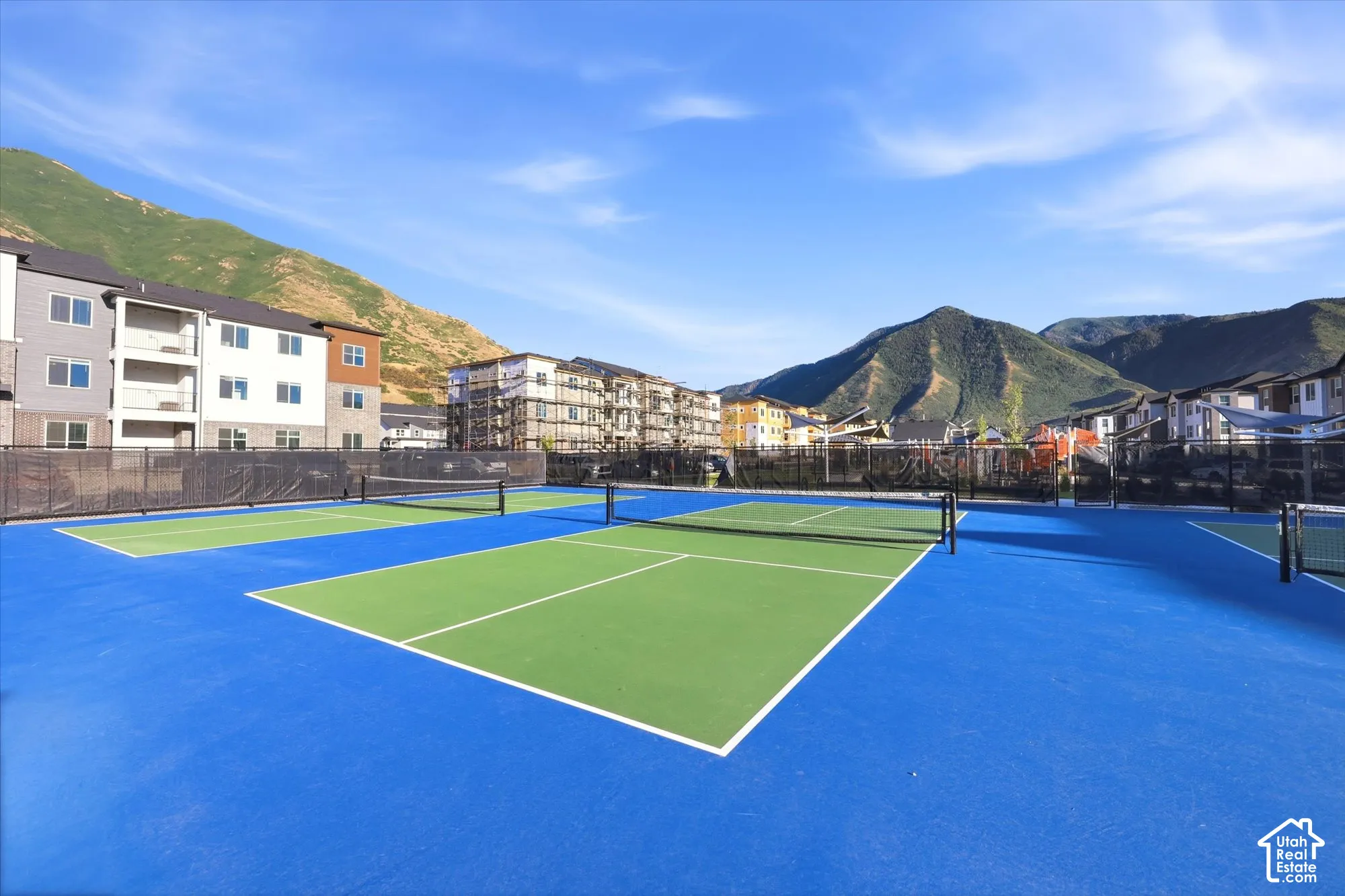 Multi-court hardscape recreation area featuring blue and green synthetic surfacing, black chain-link perimeter fencing, and mountain backdrop