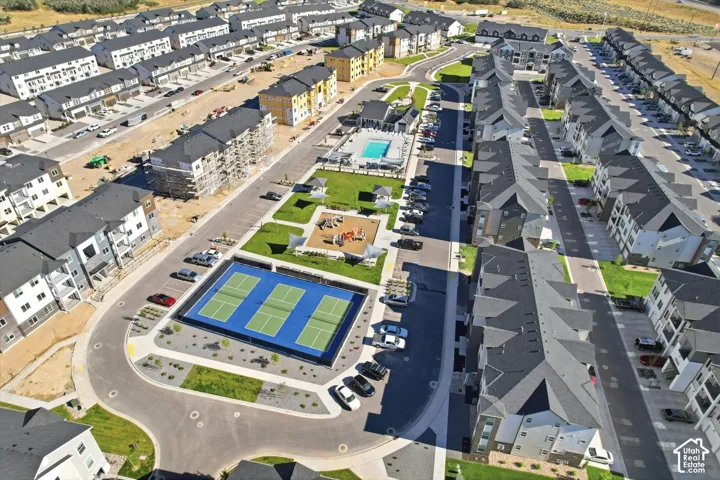 Aerial perspective of a multi-unit residential community featuring dual pickleball courts, a fenced swimming pool, a central playground, and paved surface parking