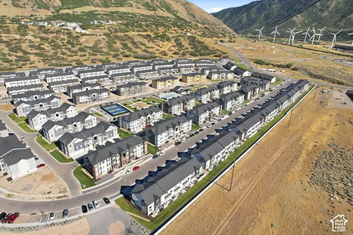 Aerial perspective of a multi-unit residential community featuring asphalt shingle roofing, paved roadways, manicured lawn areas, a fenced tennis court, and mountain terrain backdrop