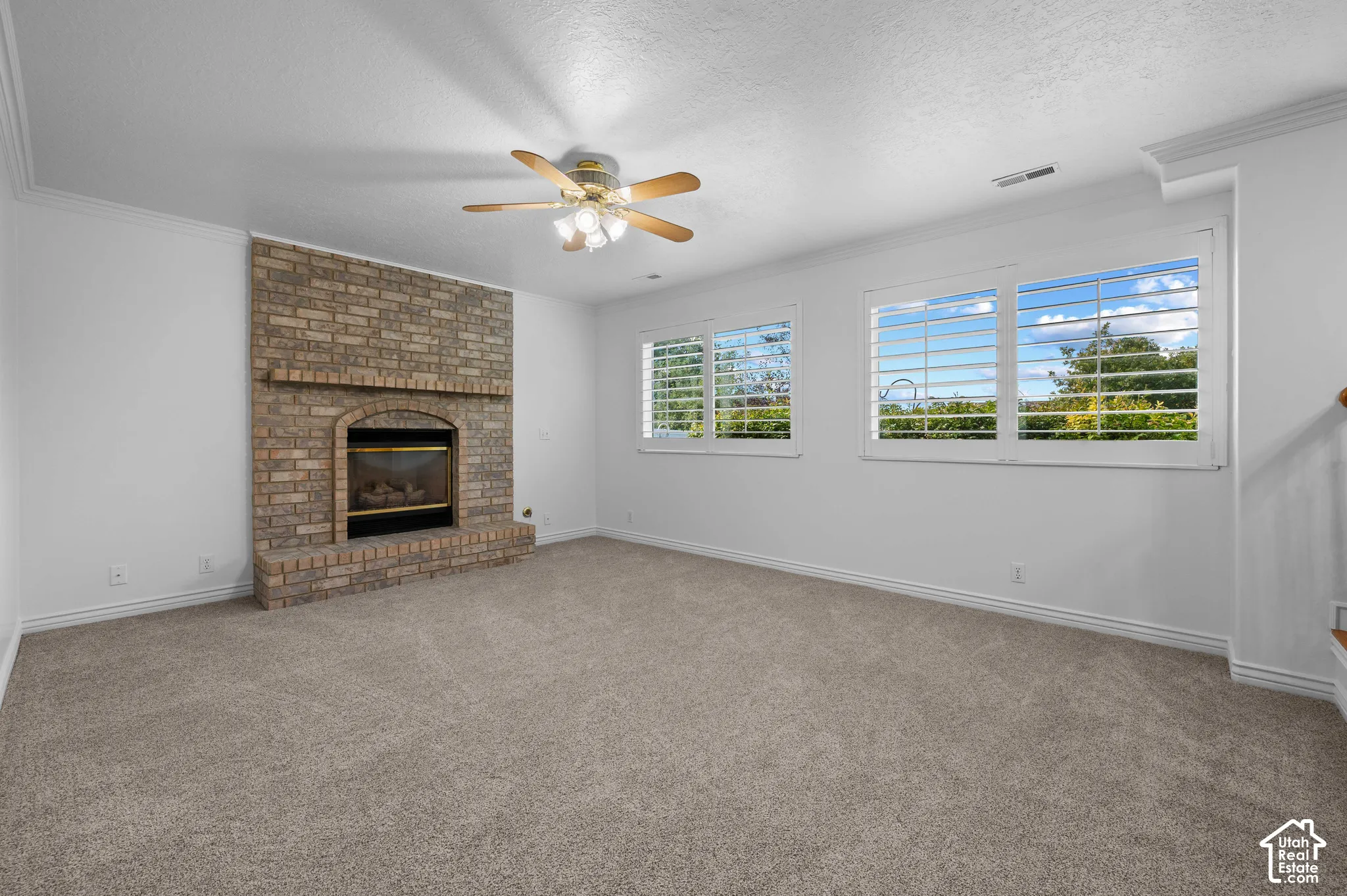 Family room with a brick fireplace with a raised hearth, plantation shutters, neutral carpeting, and a ceiling fan with multi-light fixture