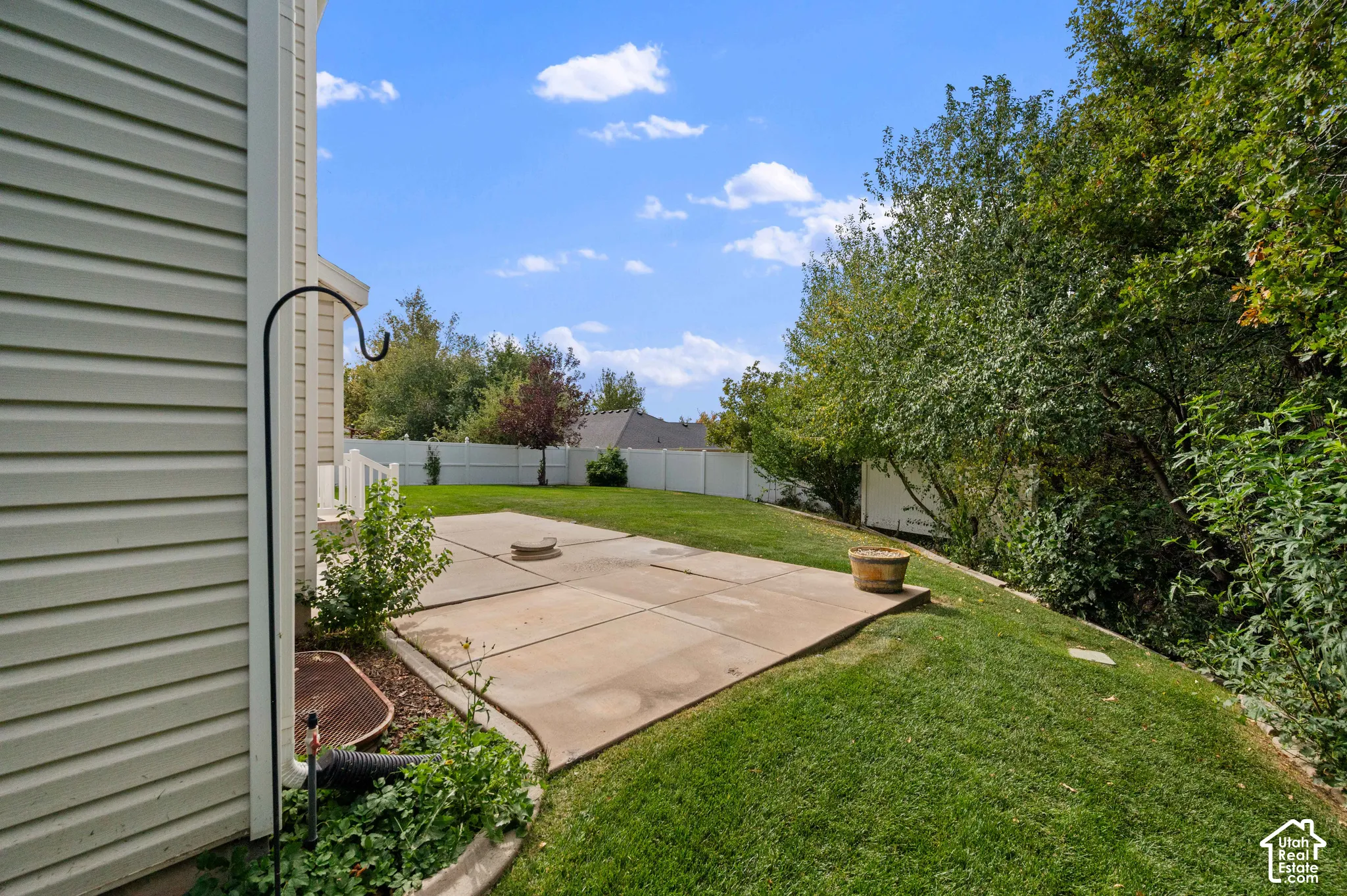 Backyard view featuring a concrete patio, manicured lawn, vinyl perimeter fencing, and mature trees