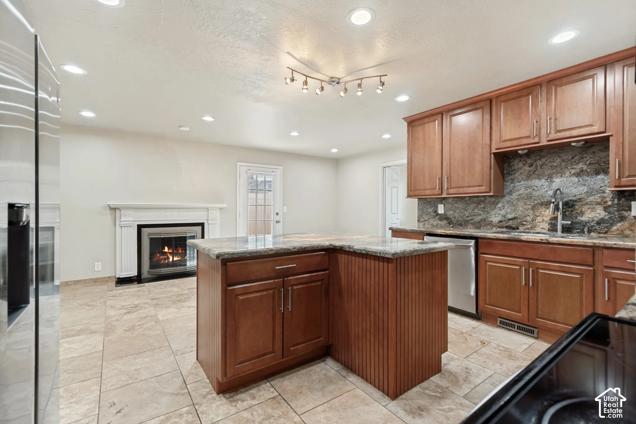 Kitchen featuring wood cabinetry, stone-surface countertops, stone-accented backsplash, central kitchen island, and recessed ceiling lighting, complemented by tile flooring and a built-in fireplace