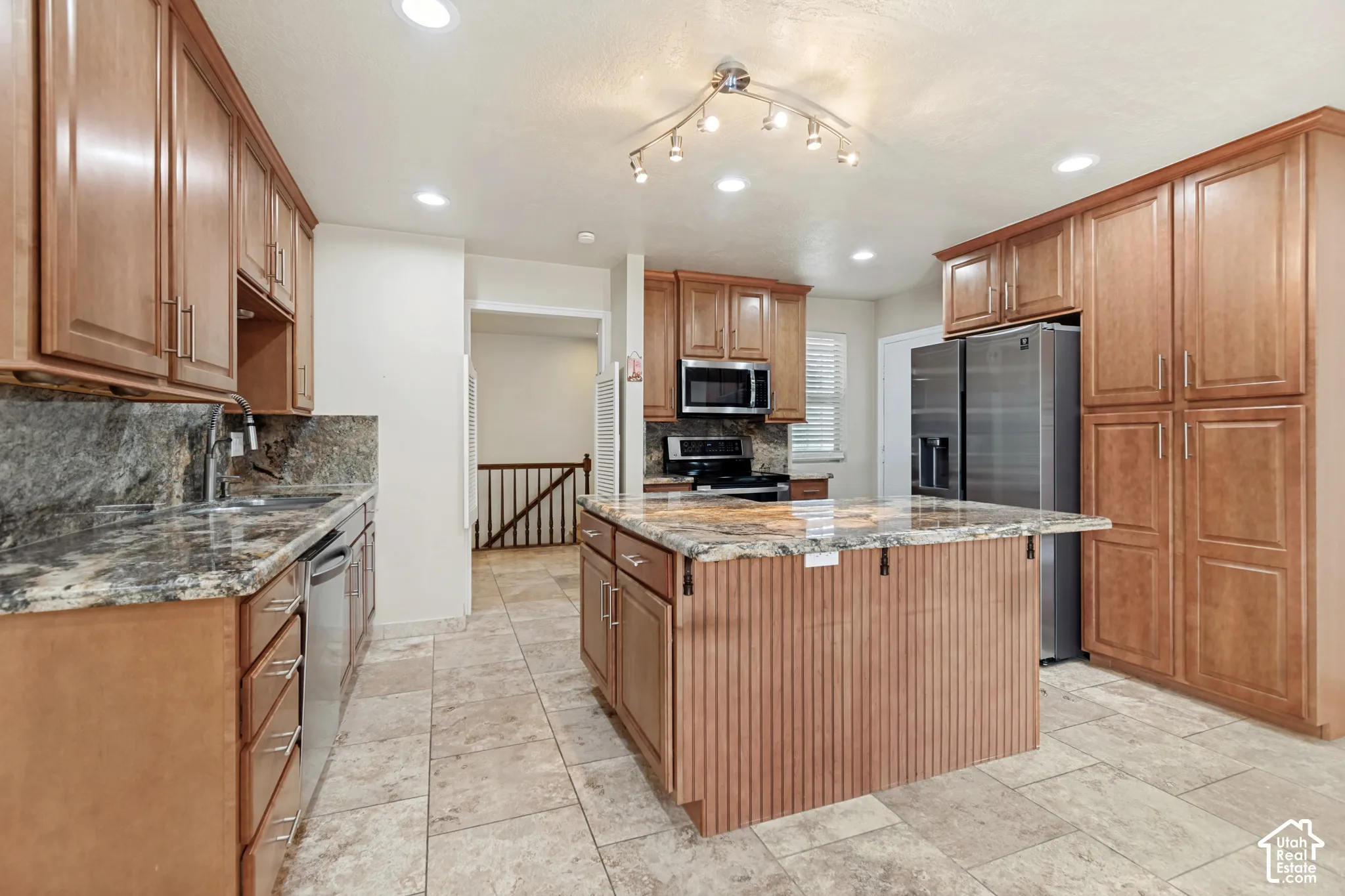 Kitchen featuring natural wood cabinetry, stone-patterned countertops, stainless steel appliances, a central island with beadboard detailing, and stone-finish floor tile