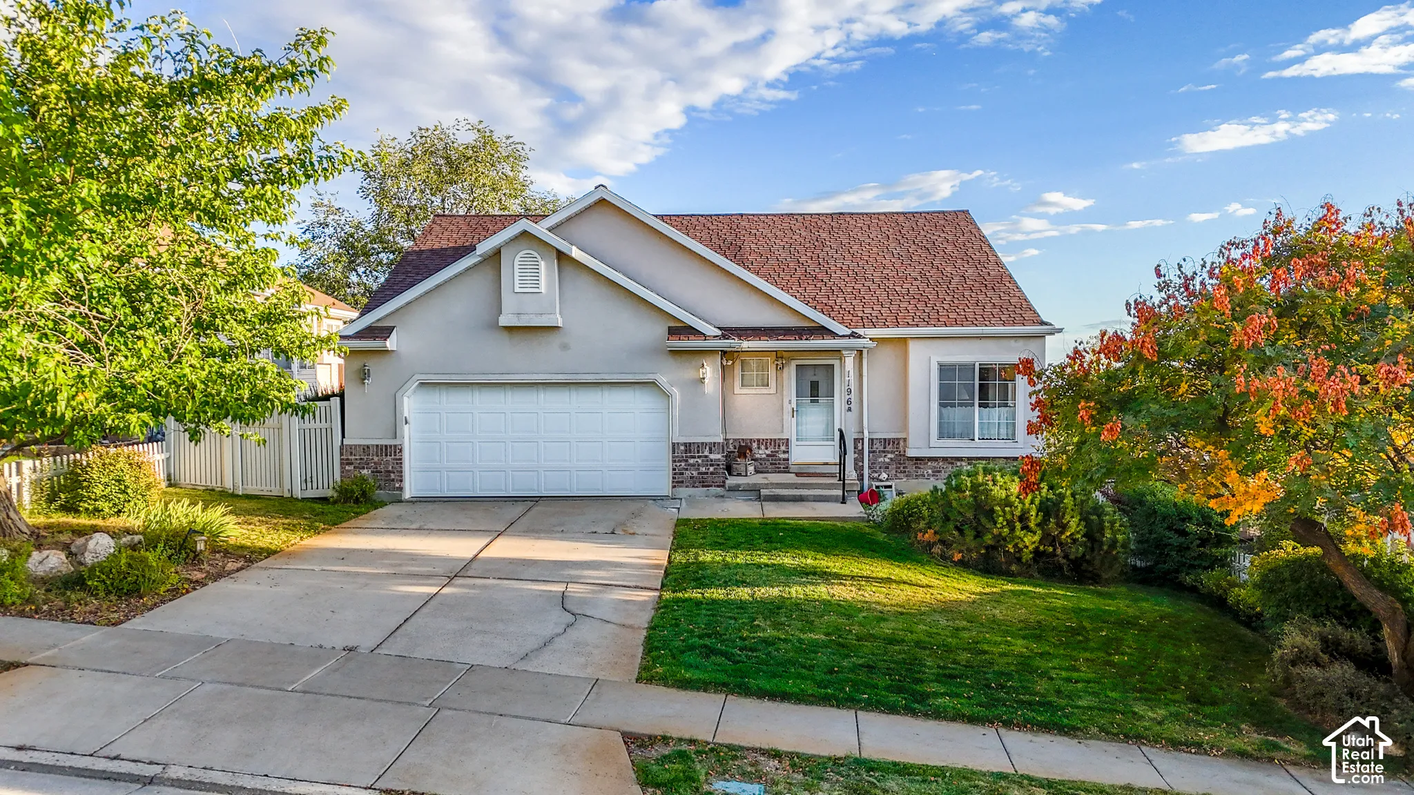 Single-story exterior featuring light-toned stucco siding, brick masonry detailing, an asphalt shingle roof, a front-facing attached two-car garage, and concrete driveway
