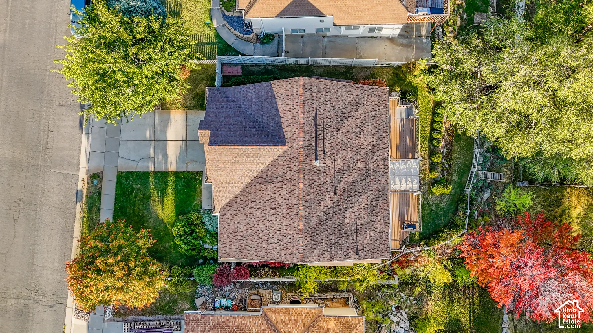 Aerial perspective of a property featuring an expansive rear wood deck, a concrete driveway, multi-level tiered landscaping, and mature perimeter trees