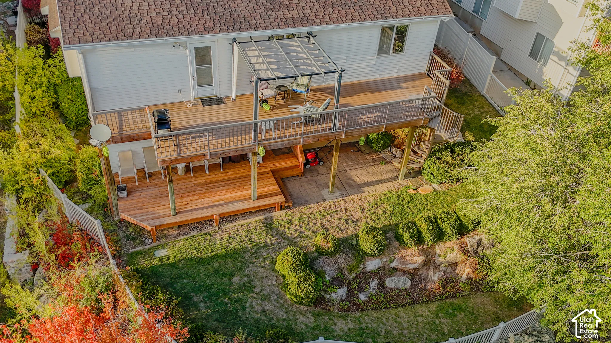 Multi-level elevated wood deck featuring a metal railing, pergola, lower-level wood patio, and stone retaining wall with mature landscaping