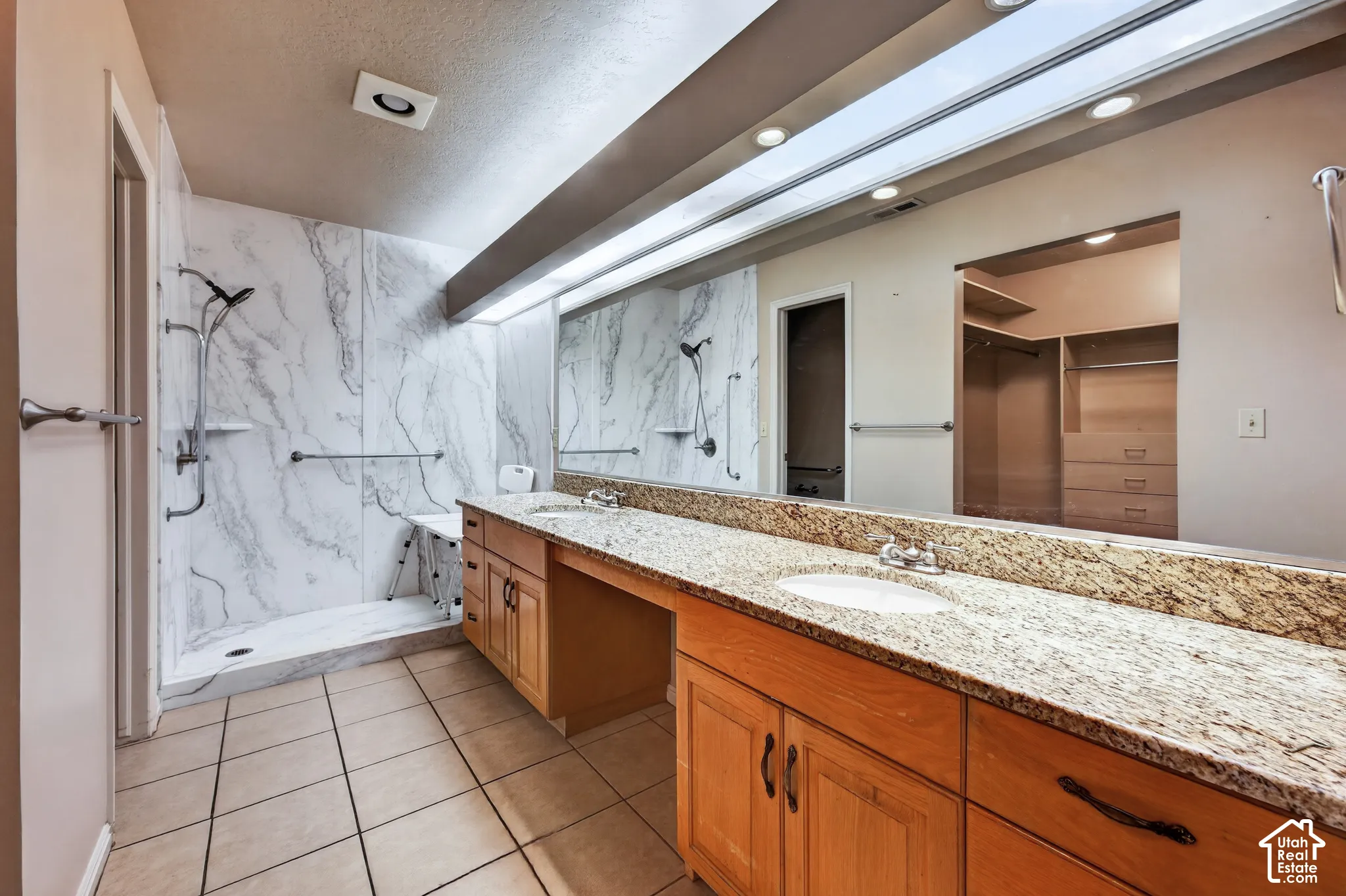Dual-sink vanity with stone countertop and wood cabinetry, expansive full-wall mirror, walk-in shower with stone-patterned surround, and tile flooring