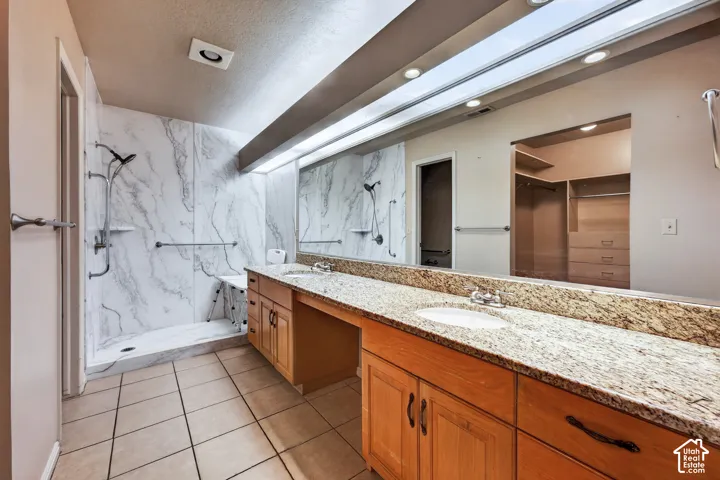 Dual-sink vanity with stone countertop and wood cabinetry, expansive full-wall mirror, walk-in shower with stone-patterned surround, and tile flooring