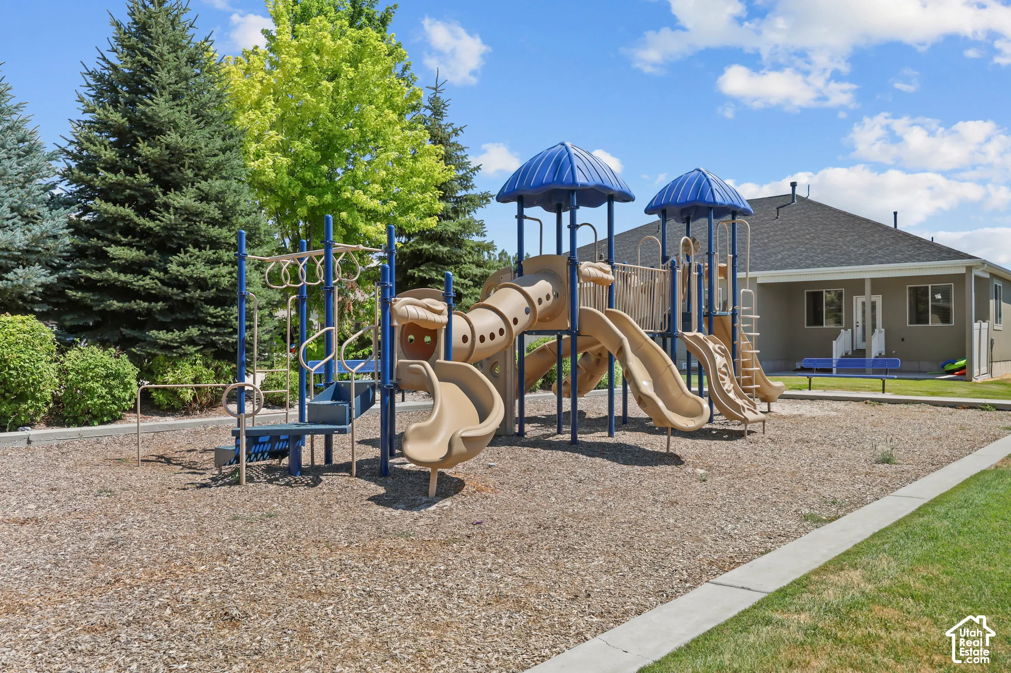 Recreational area featuring a multi-component play structure with slides and climbing apparatus, set within a graveled surface, bordered by manicured lawn and mature coniferous trees